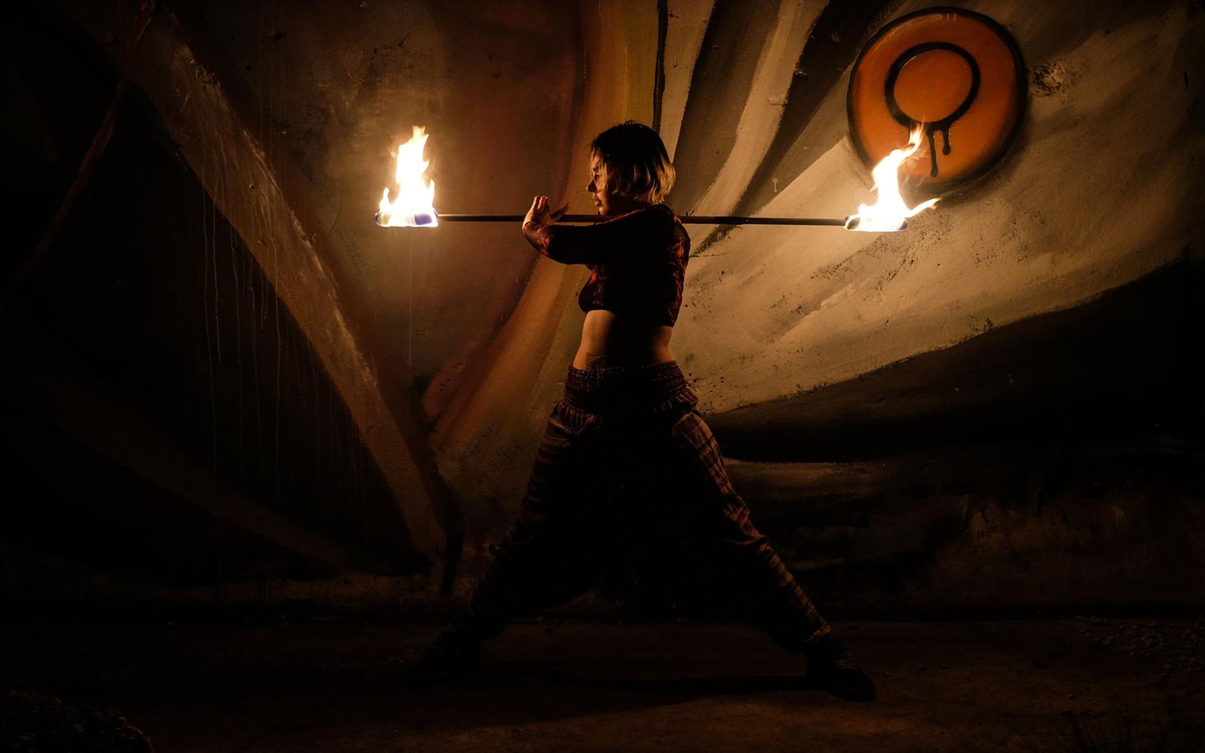 Wide shot of a performer in a wide stance holding a burning staff in front of a mural with a round emblem