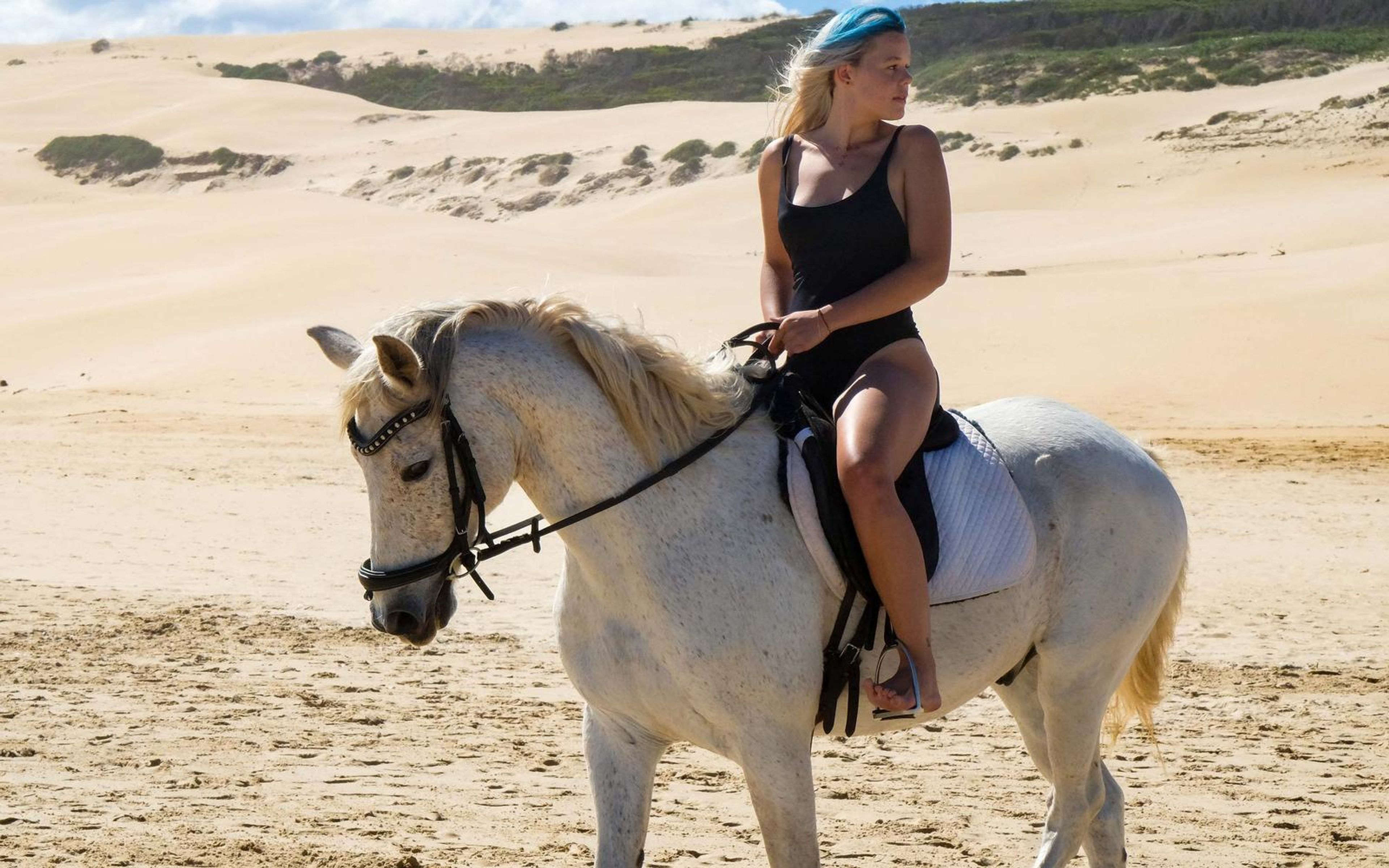 Woman in a black swimsuit riding a white horse across a sandy beach, looking off to the side