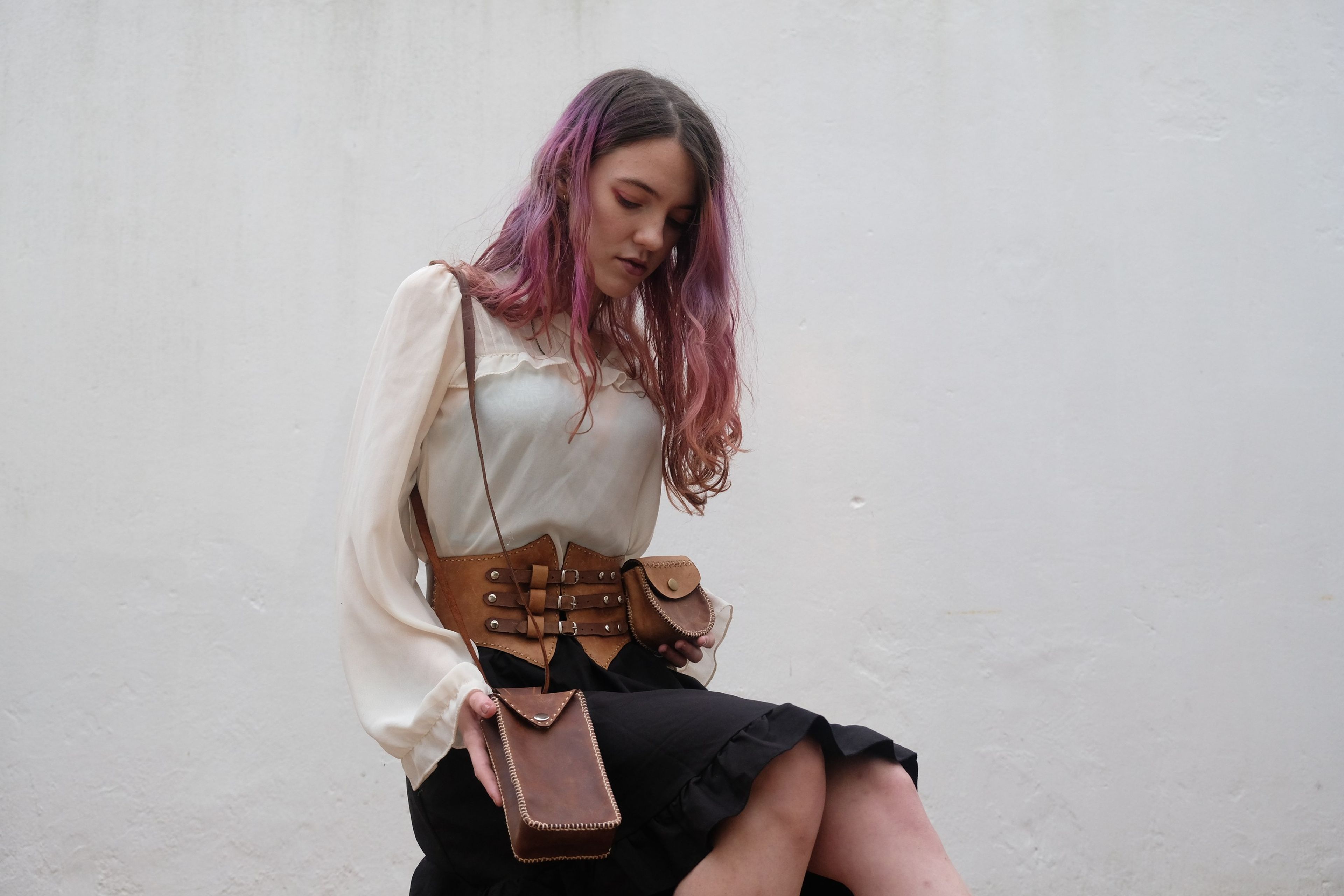 Woman with pink-purple hair looking down, wearing a leather corset belt and carrying a brown bag