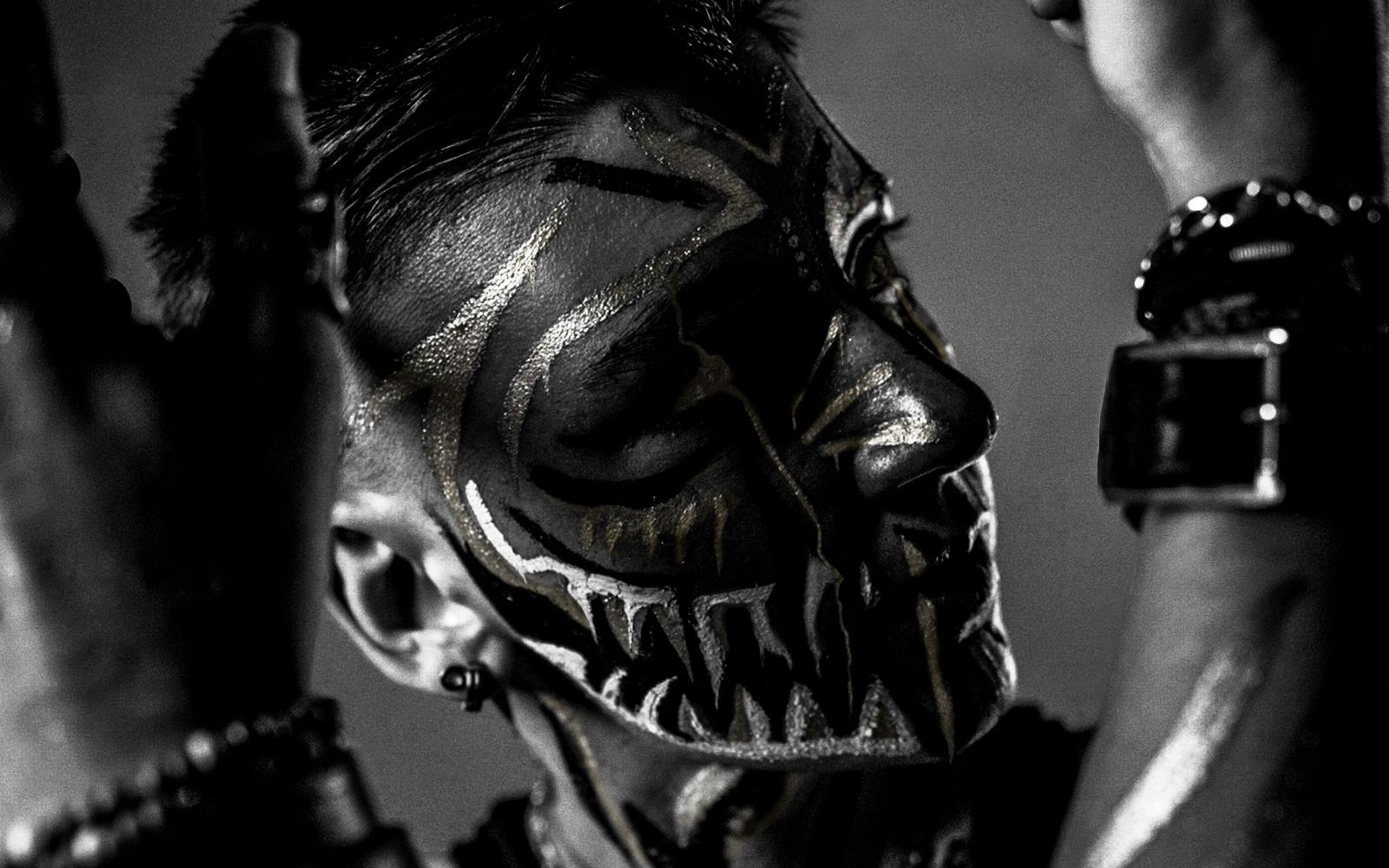 Black and white portrait of a performer with fang-like face paint, hands raised beside the head, studded wristbands