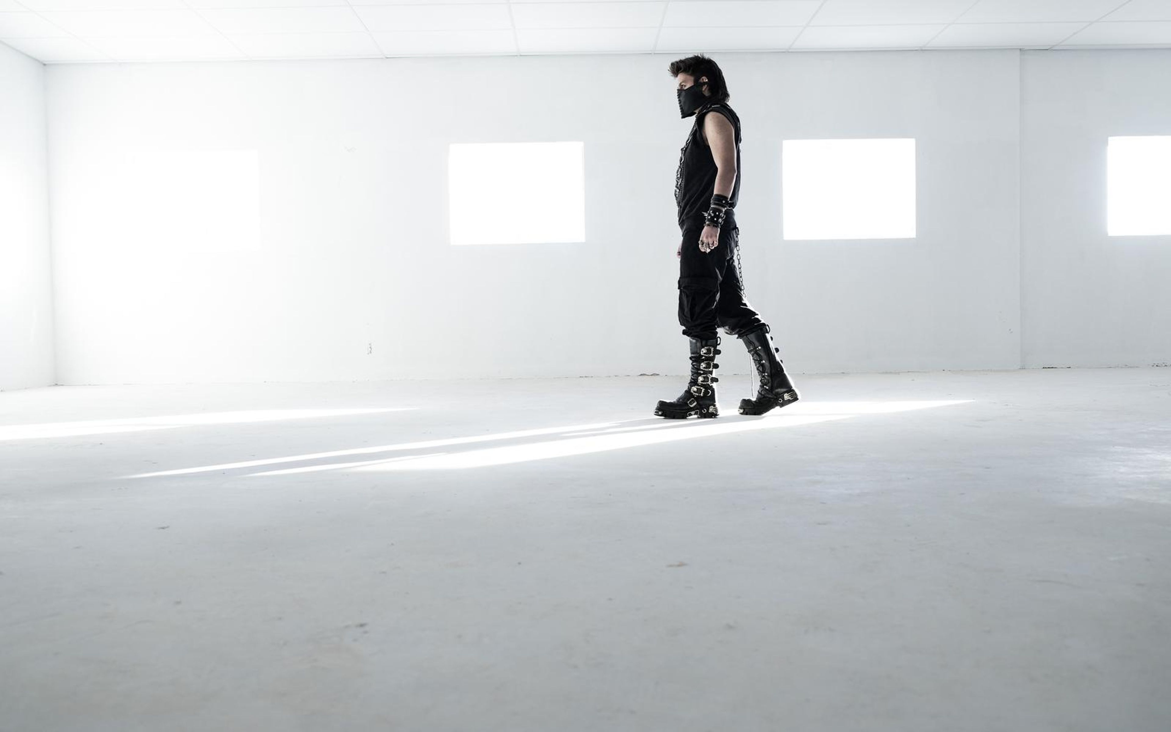 Man in black and buckled boots walking in profile across a pale, overexposed white room with light streaks on the floor