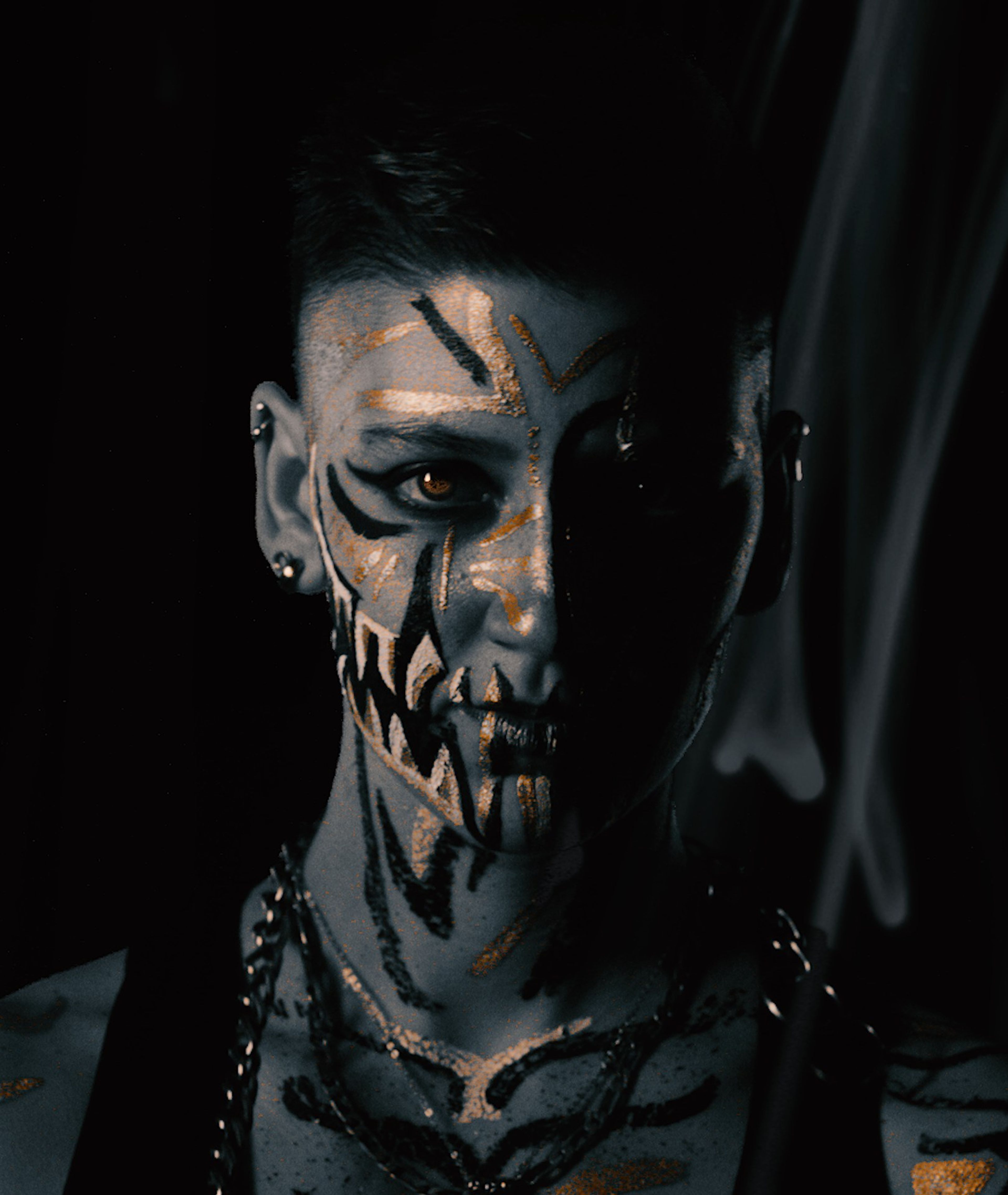 Face-painted portrait with black fang markings and gold highlights, lit from the front in deep shadow