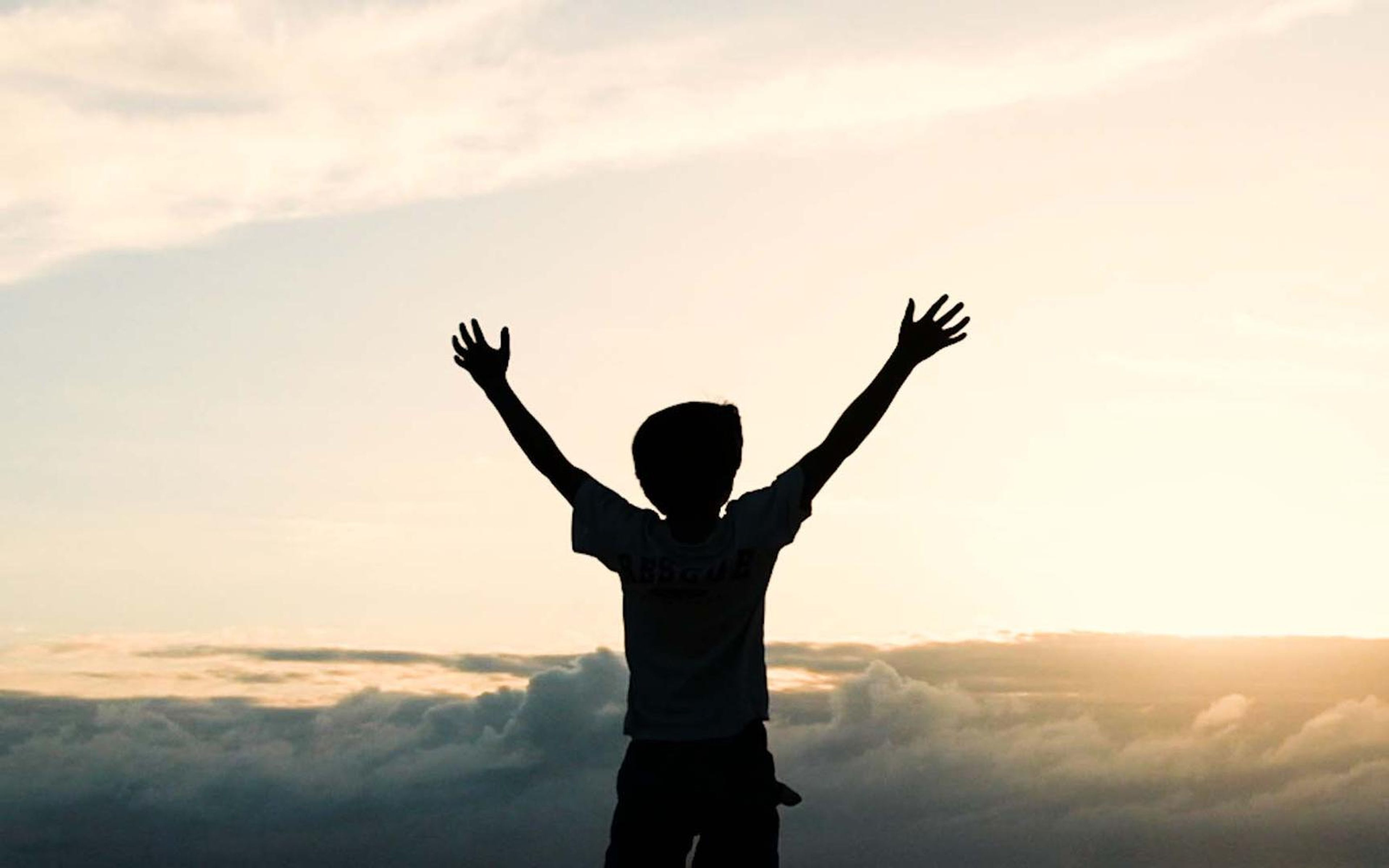 Silhouette of a child with arms raised against a golden sky above a layer of clouds