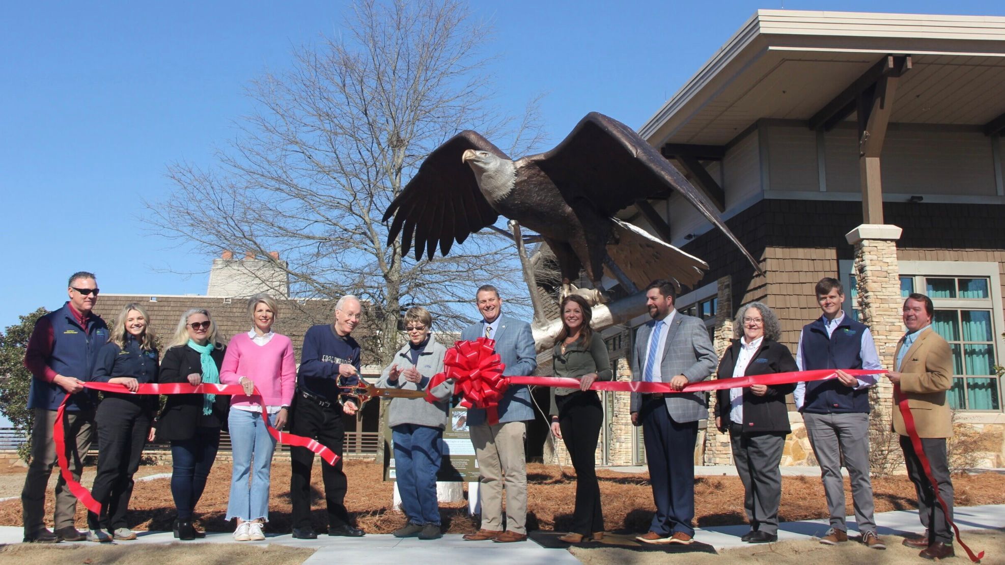 This is a statue unveiling at Lake Guntersville State Park. A group of people stand in front of a building, cutting a red ribbon with oversized scissors.