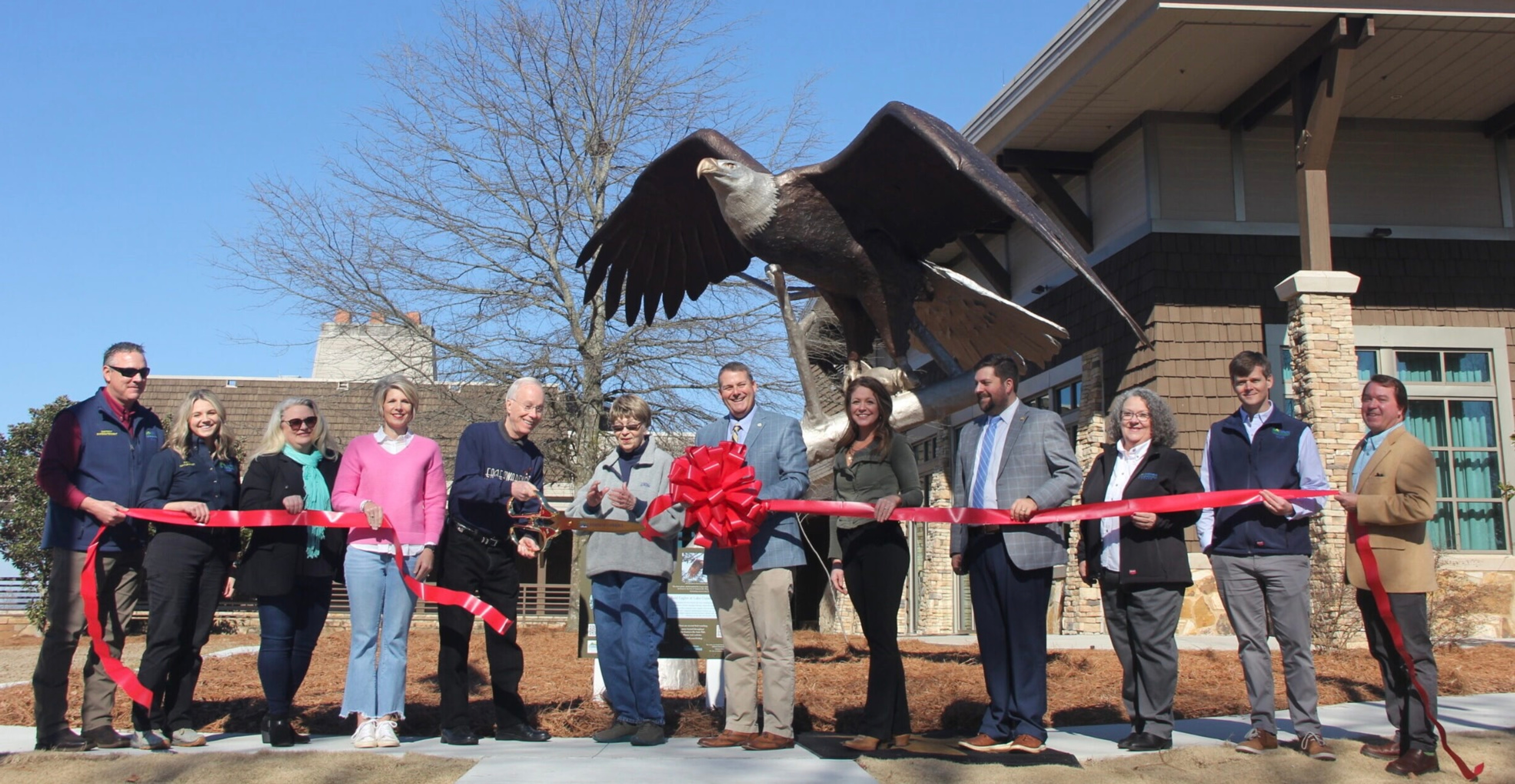 This is a statue unveiling at Lake Guntersville State Park. A group of people stand in front of a building, cutting a red ribbon with oversized scissors.