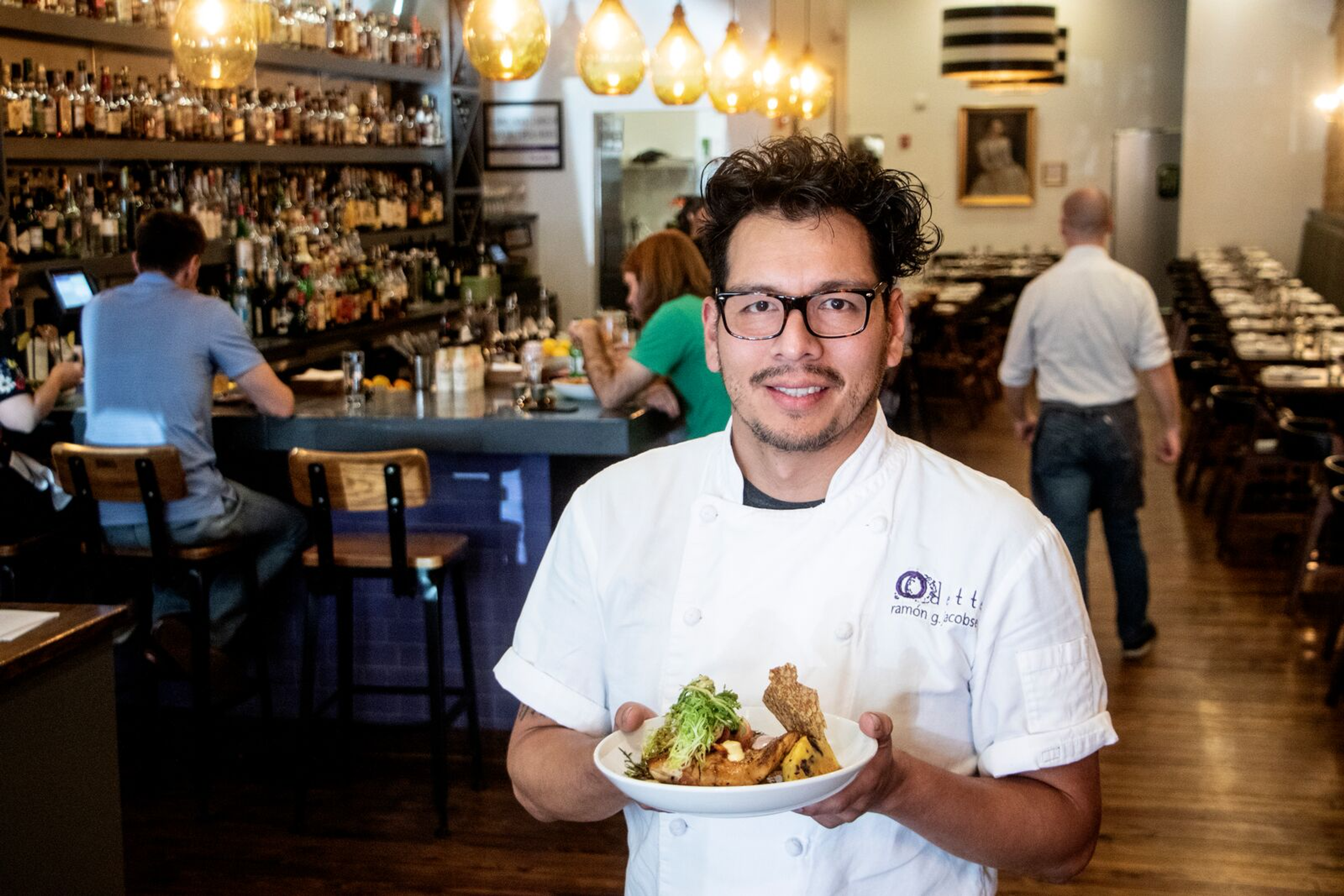 A chef in a white uniform holds a plated dish posing for a photo in a restaurant. Behind him is a bar with people seated, and more tables with people in the background.