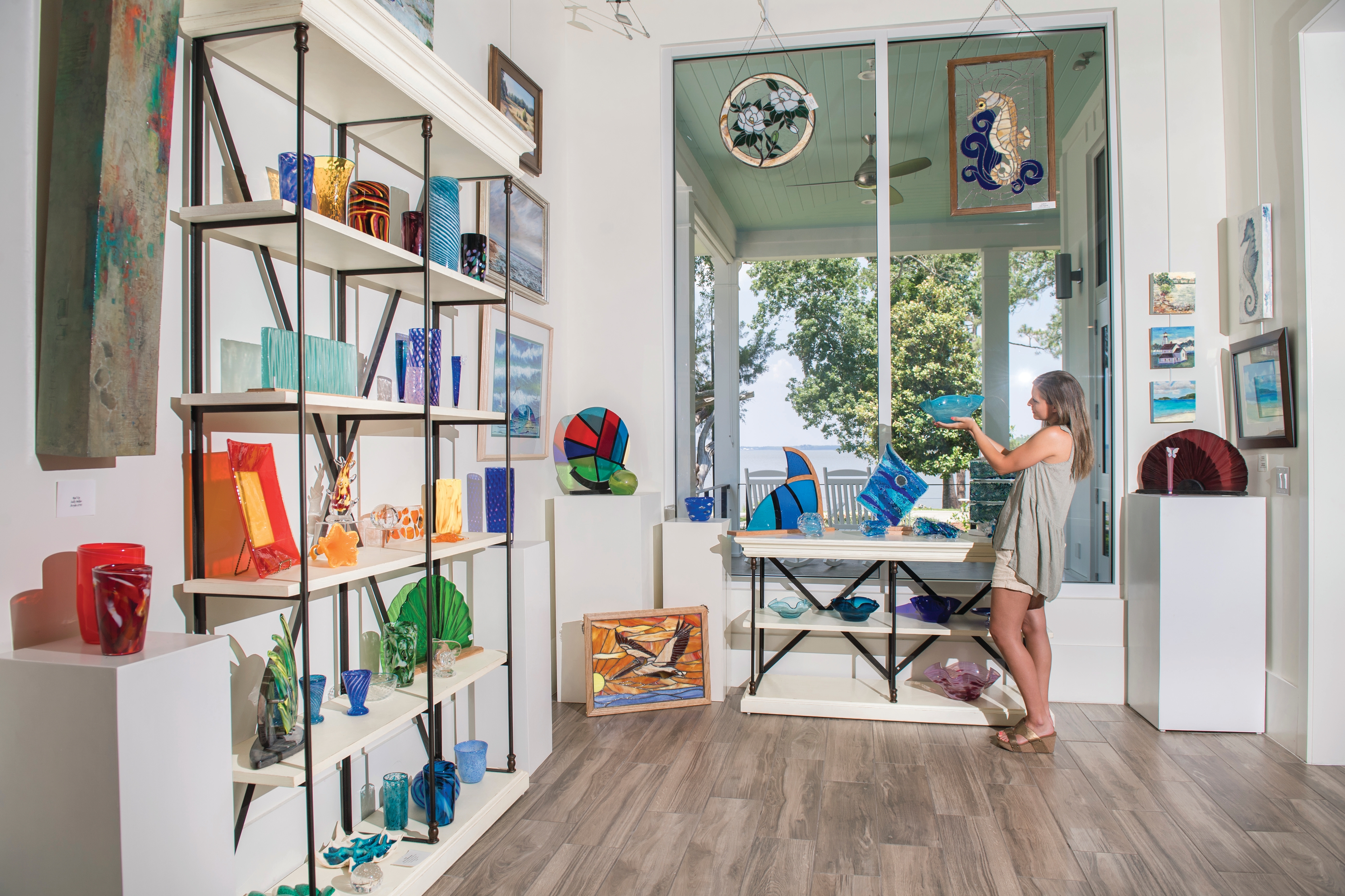 A woman at Orange Beach Coastal Arts Center stands in a bright art gallery and is looking at a piece of stained glass on display.