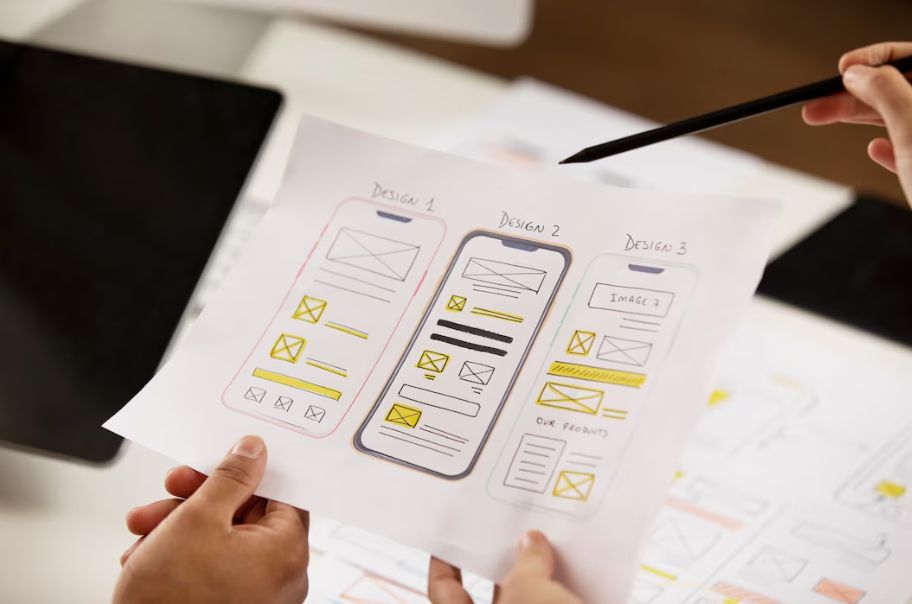 Hands holding a paper with sketches of three different mobile app designs labeled Design 1, Design 2, and Design 3, featuring various layouts and highlighted elements.