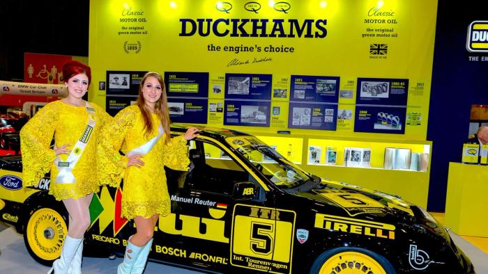 Two promoters in yellow beside a Duckhams-liveried rally car on an exhibition stand.