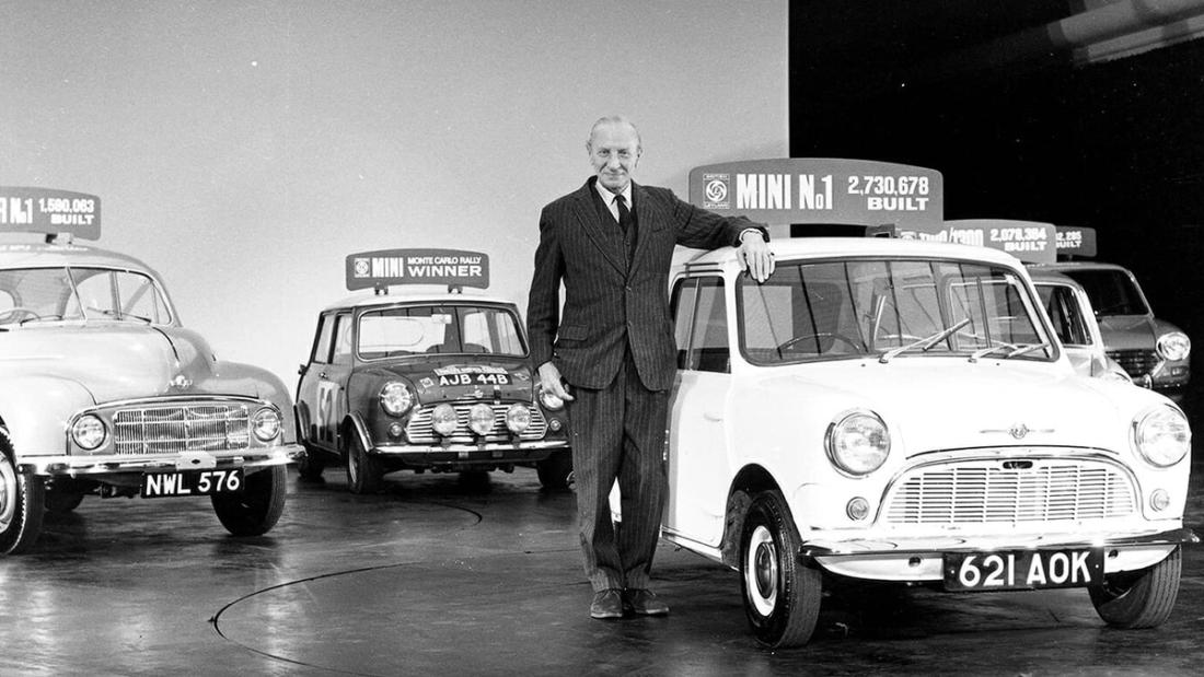 A man in a suit standing beside three early Minis lined up indoors.