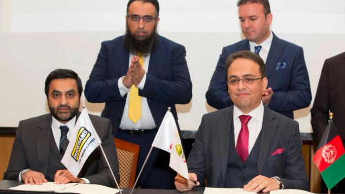 Two men signing an agreement at a table with desk flags, colleagues applauding behind them.