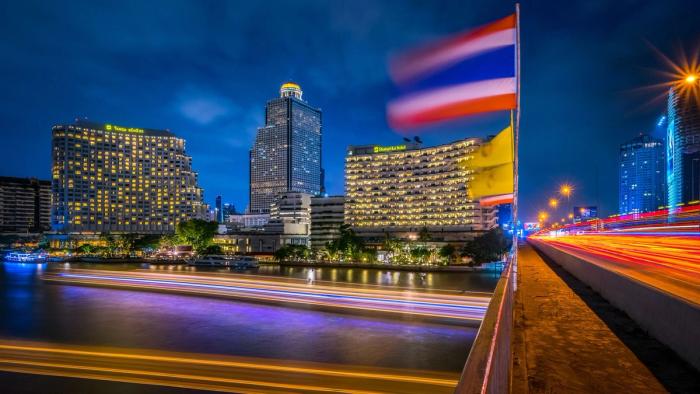 A city skyline at night with light trails along the road and a Thai flag.