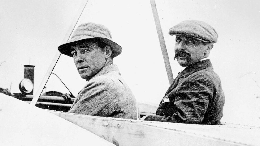Two men in flat caps and goggles seated in the open cockpit of an early biplane.