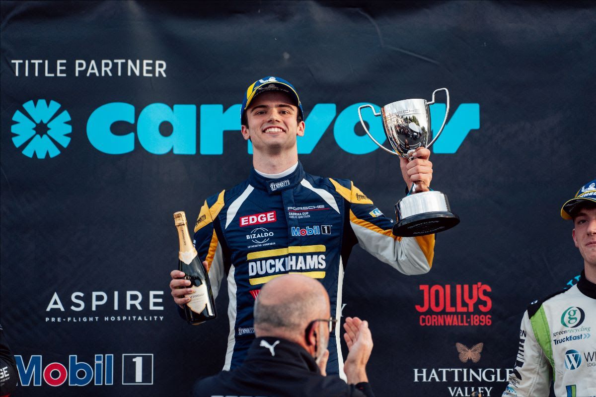 Jacob Tofts smiling on the Brands Hatch podium in his Duckhams race suit, holding up a trophy and a bottle of sparkling wine.
