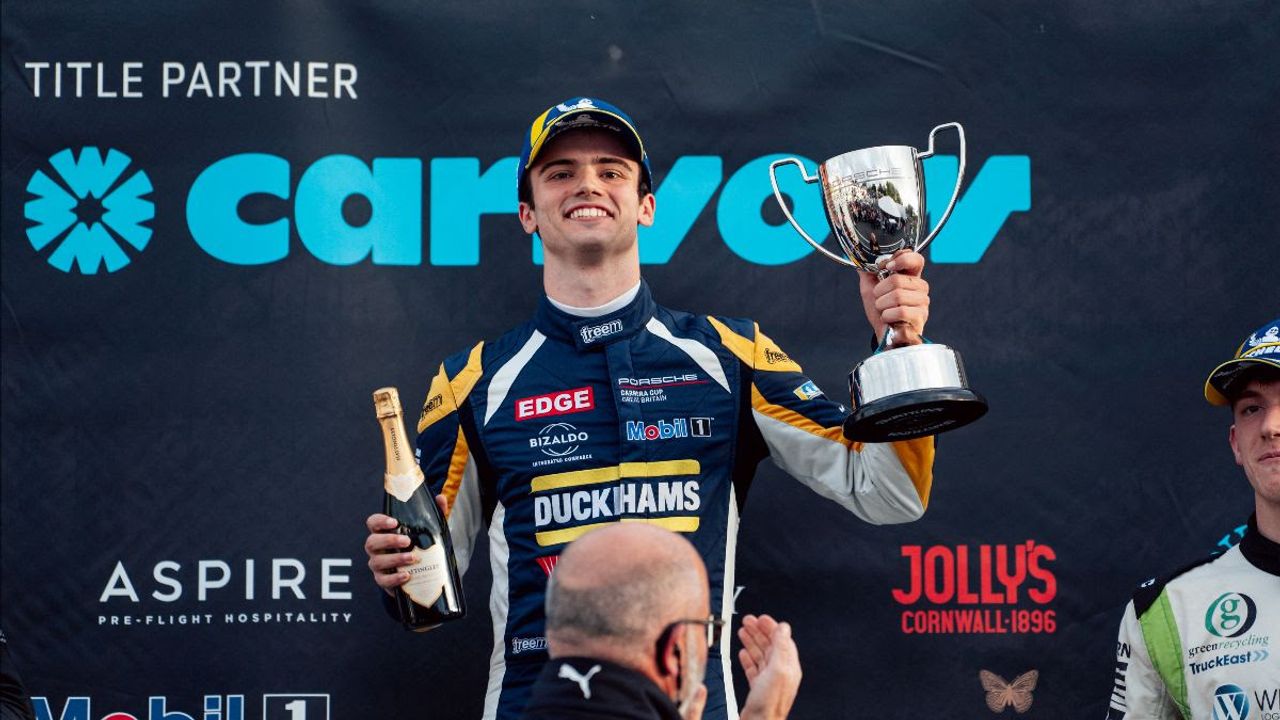 Jacob Tofts smiling on the Brands Hatch podium in his Duckhams race suit, holding up a trophy and a bottle of sparkling wine.