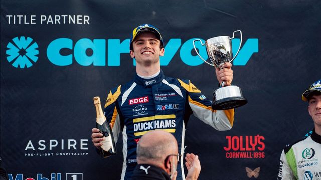 Jacob Tofts smiling on the Brands Hatch podium in his Duckhams race suit, holding up a trophy and a bottle of sparkling wine.