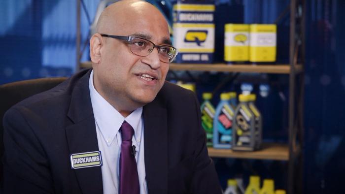 A man in a Duckhams-branded shirt and tie speaking, product shelves behind him.