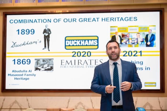 A man presenting beside a screen headed "Combination of our great heritages", pairing Duckhams in 1899 with Al Masaood in 1869.