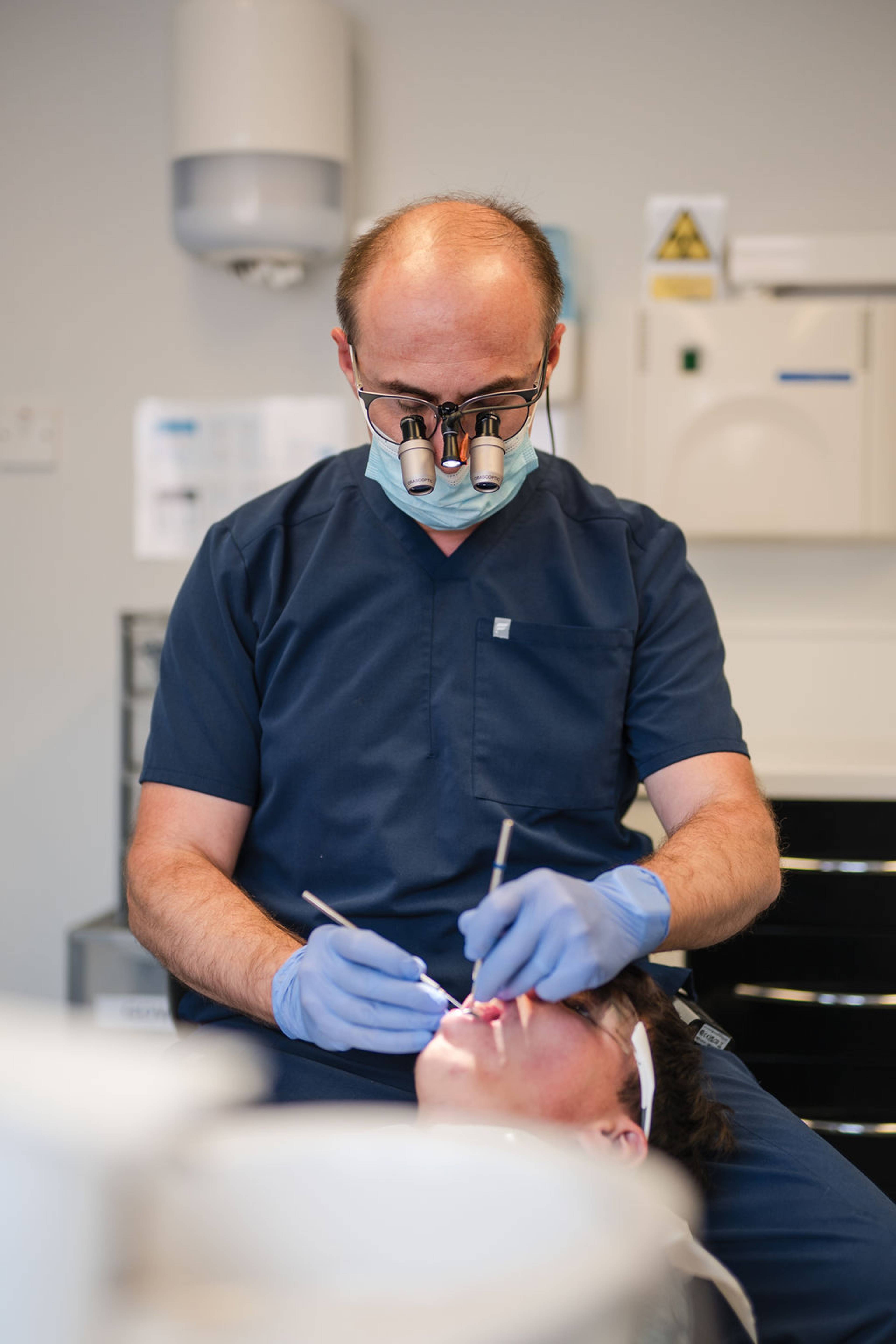 Dental professional wearing magnification loupes and protective equipment performing an oral examination on a patient in a dental treatment room.