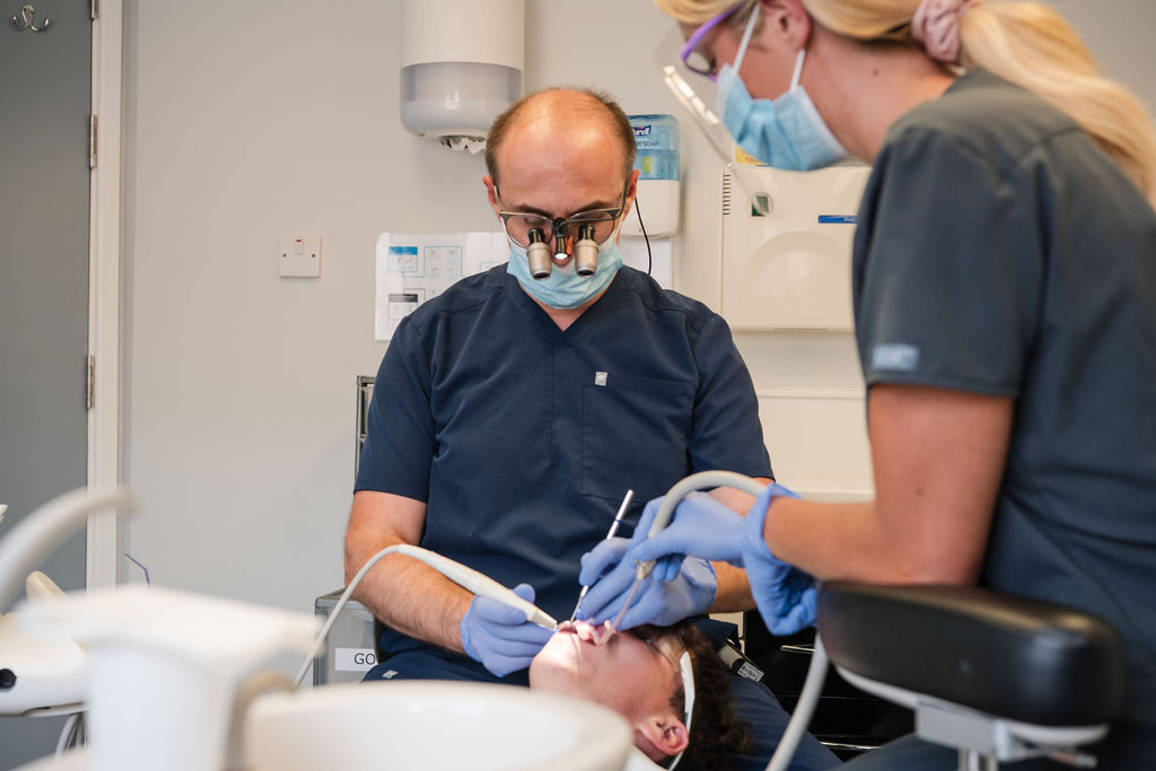 Dental professionals performing a dental procedure on a patient in a treatment room using magnification loupes, dental instruments, and suction equipment.