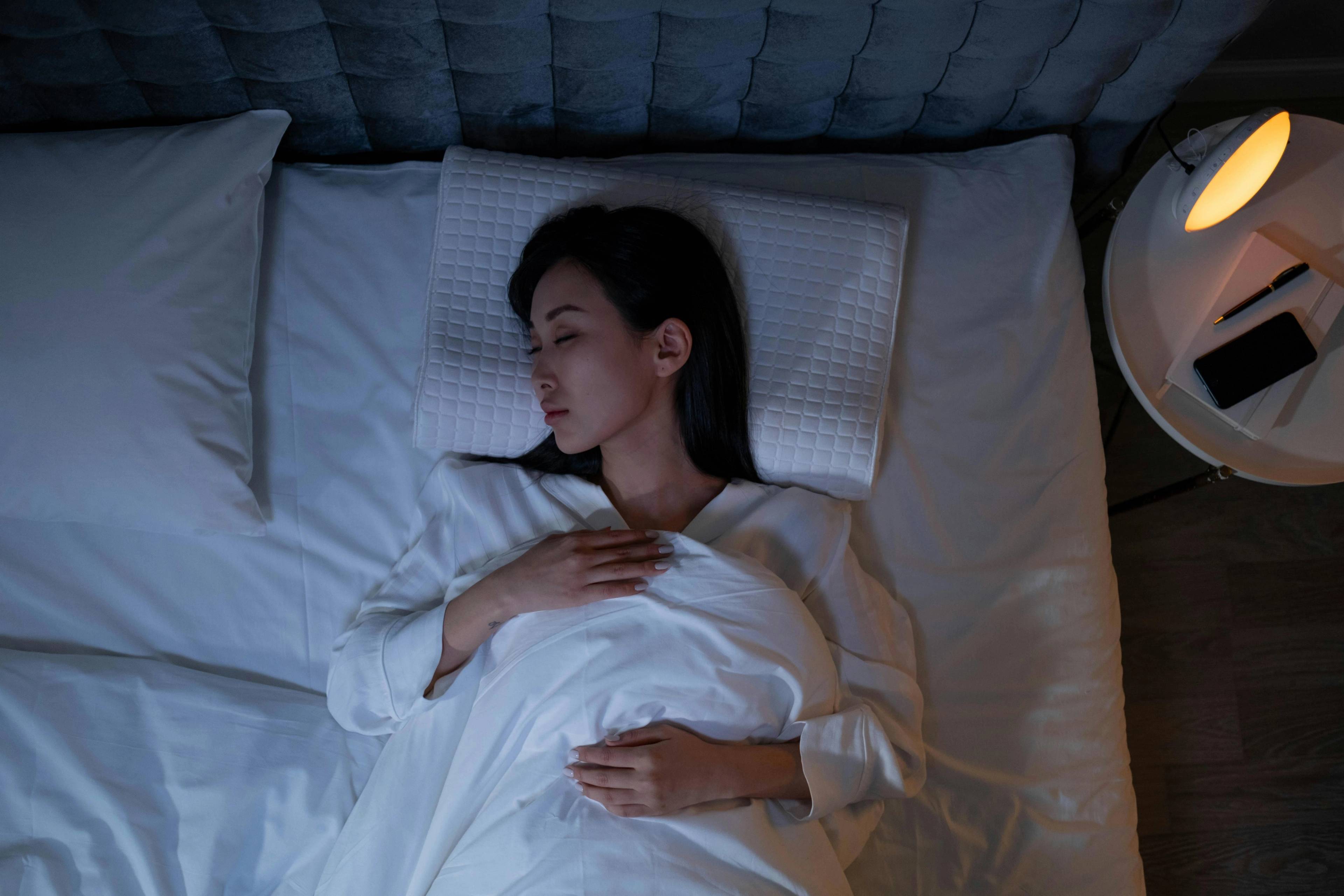 Overhead view of a person lying in bed under a white duvet in a dimly lit bedroom. The bed has white pillows and a padded blue headboard, while a bedside table holds a lit lamp, a smartphone, and a pen. Soft blue and warm yellow lighting create a nighttime setting.