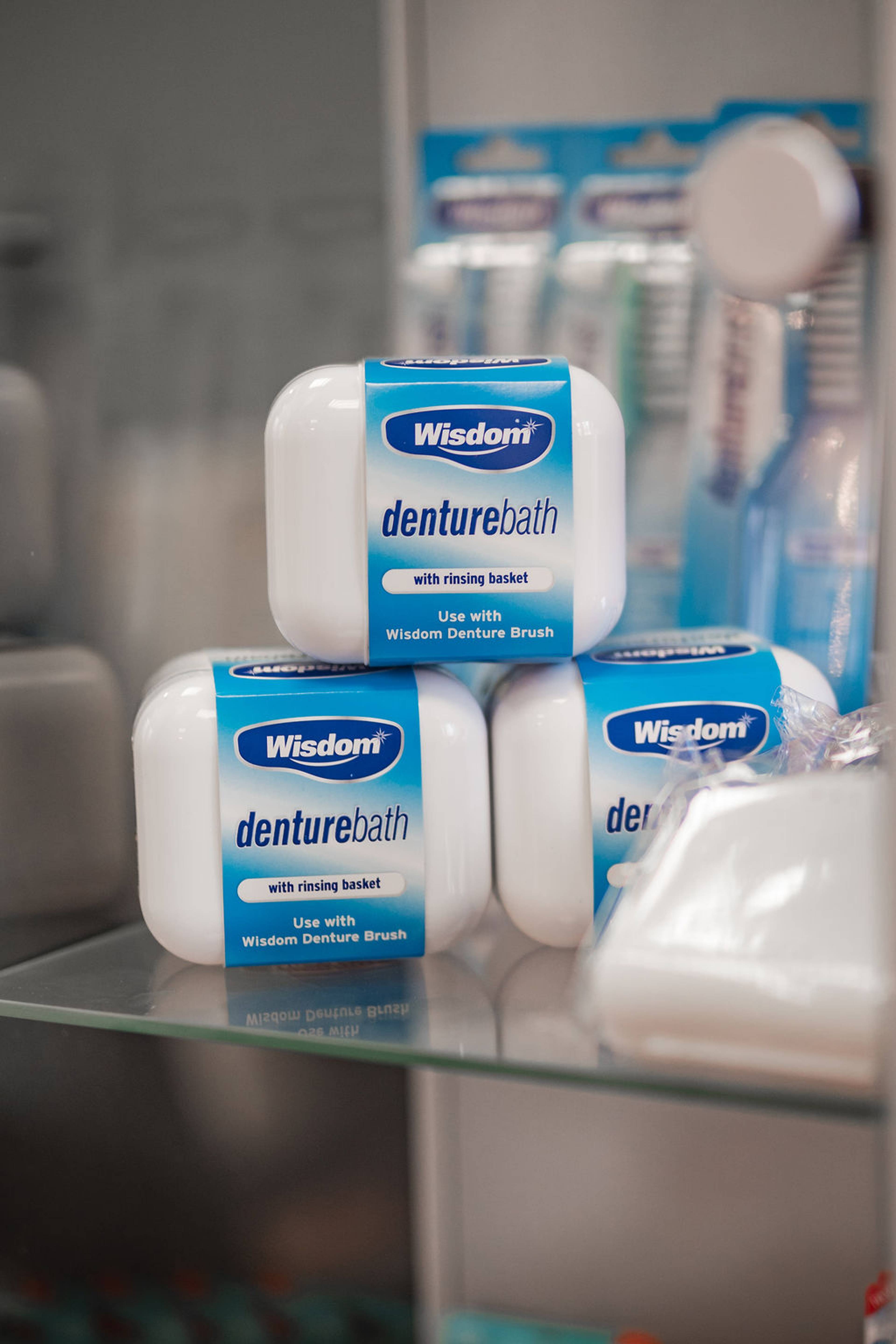 Denture care products displayed on a glass shelf, including Wisdom Denture Bath containers with rinsing baskets.
