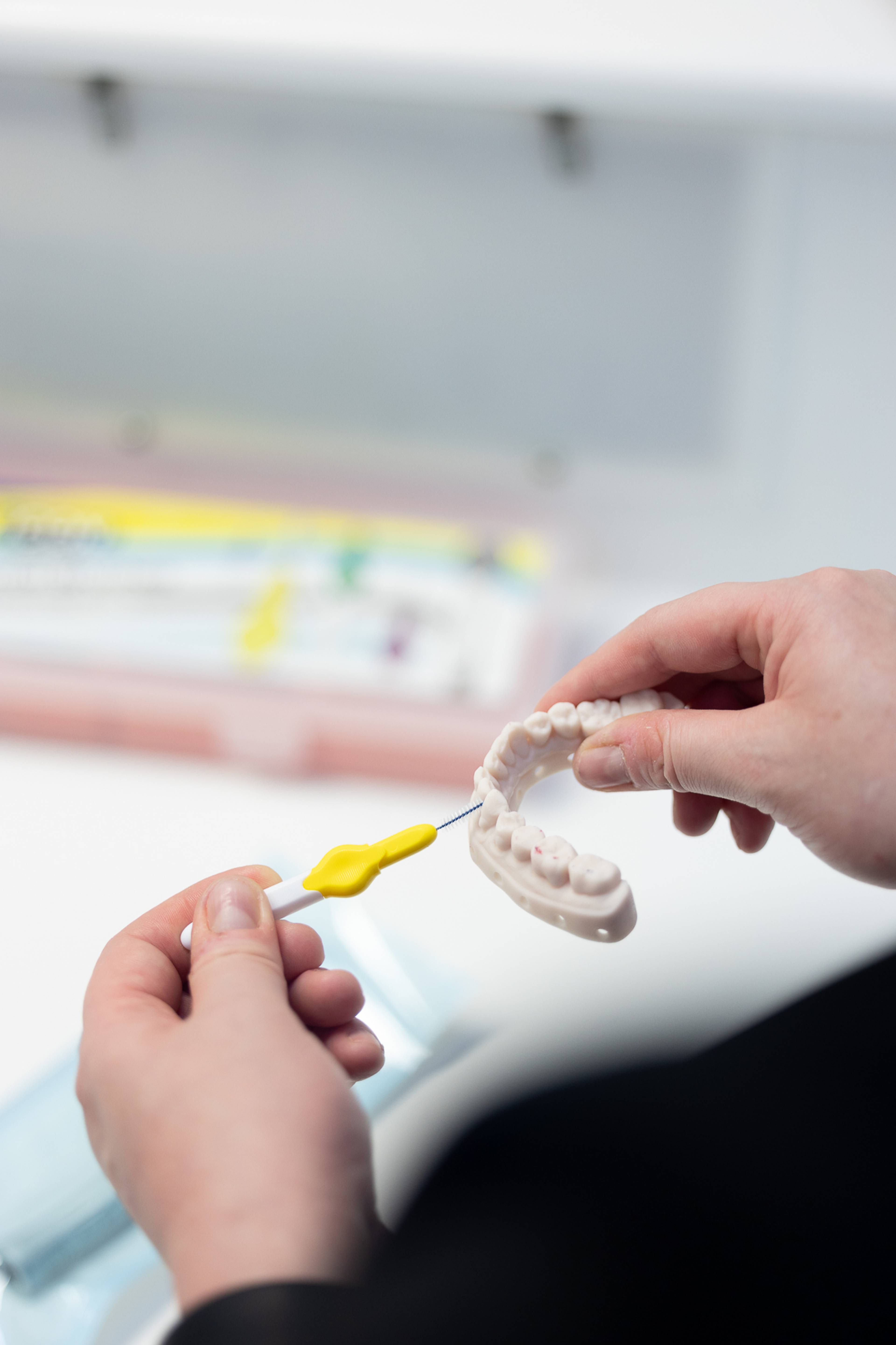 Close-up of two hands holding a dental model shaped like a lower jaw while using a small interdental brush with a yellow handle to clean between the model teeth. The background is softly blurred, showing a clinical dental setting with dental care supplies and equipment out of focus.
