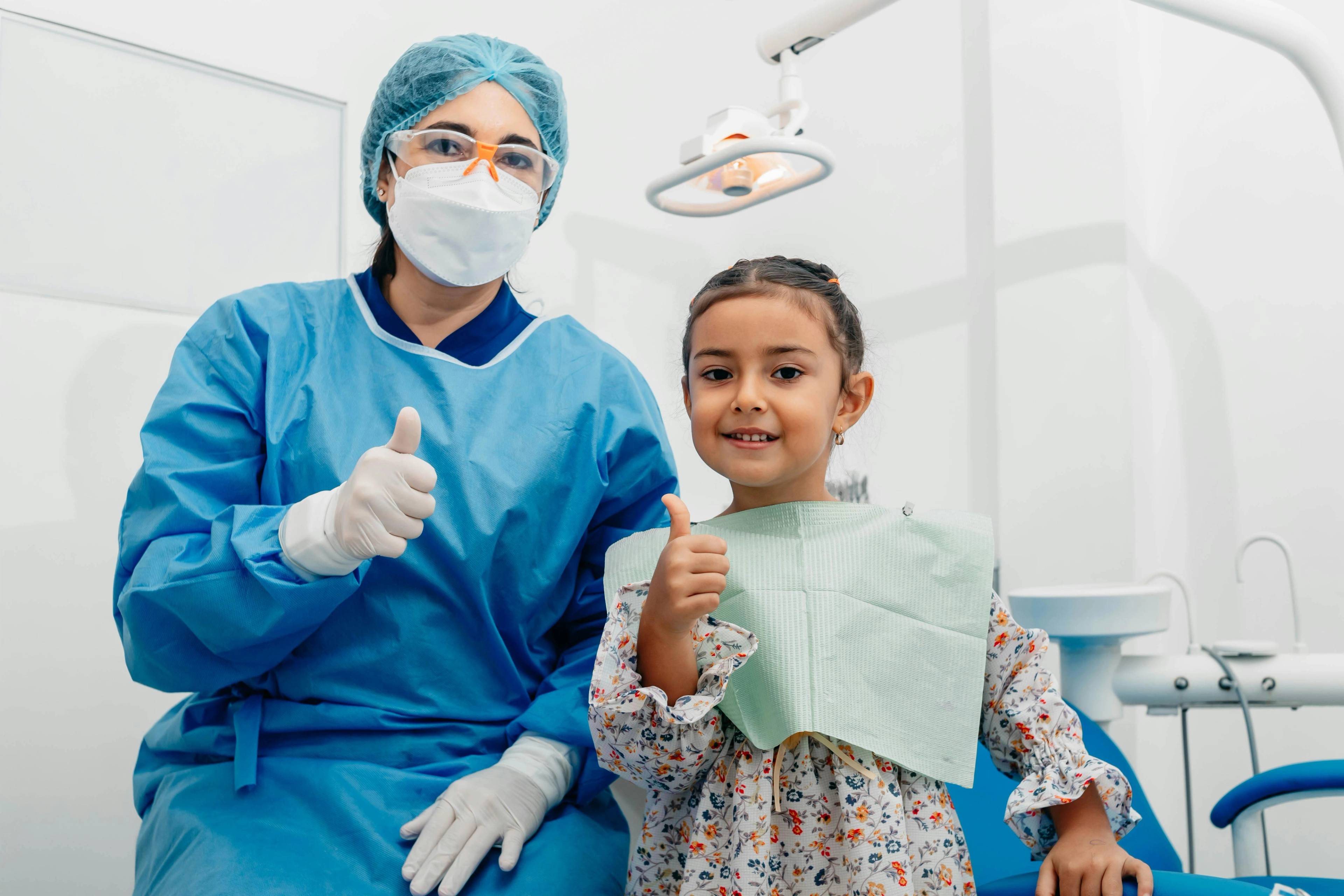 Child seated in a dental chair and dental professional in protective clothing both giving a thumbs-up in a dental treatment room.