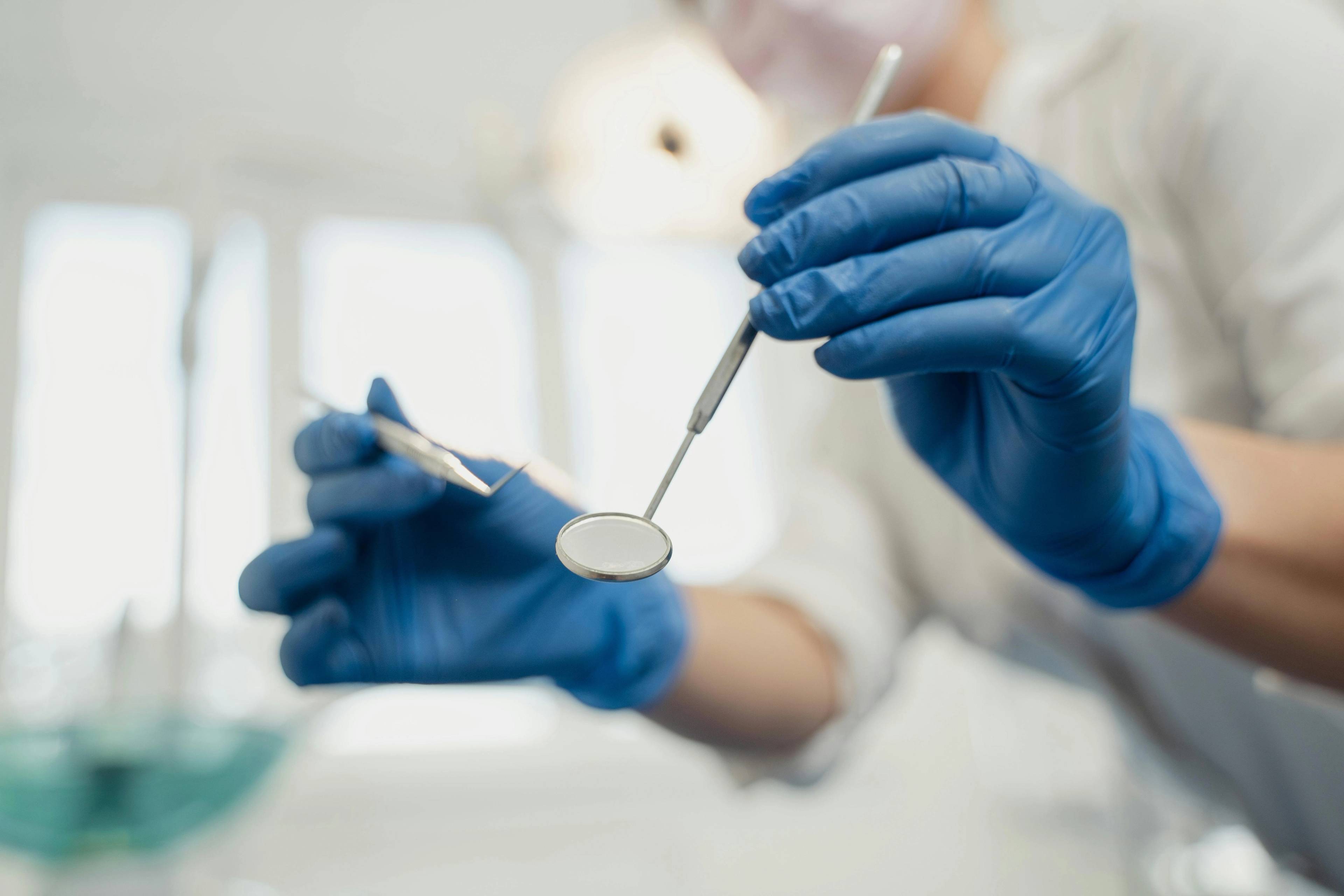 Close-up of gloved hands holding a dental mirror and dental probe during an oral examination in a clinical setting.