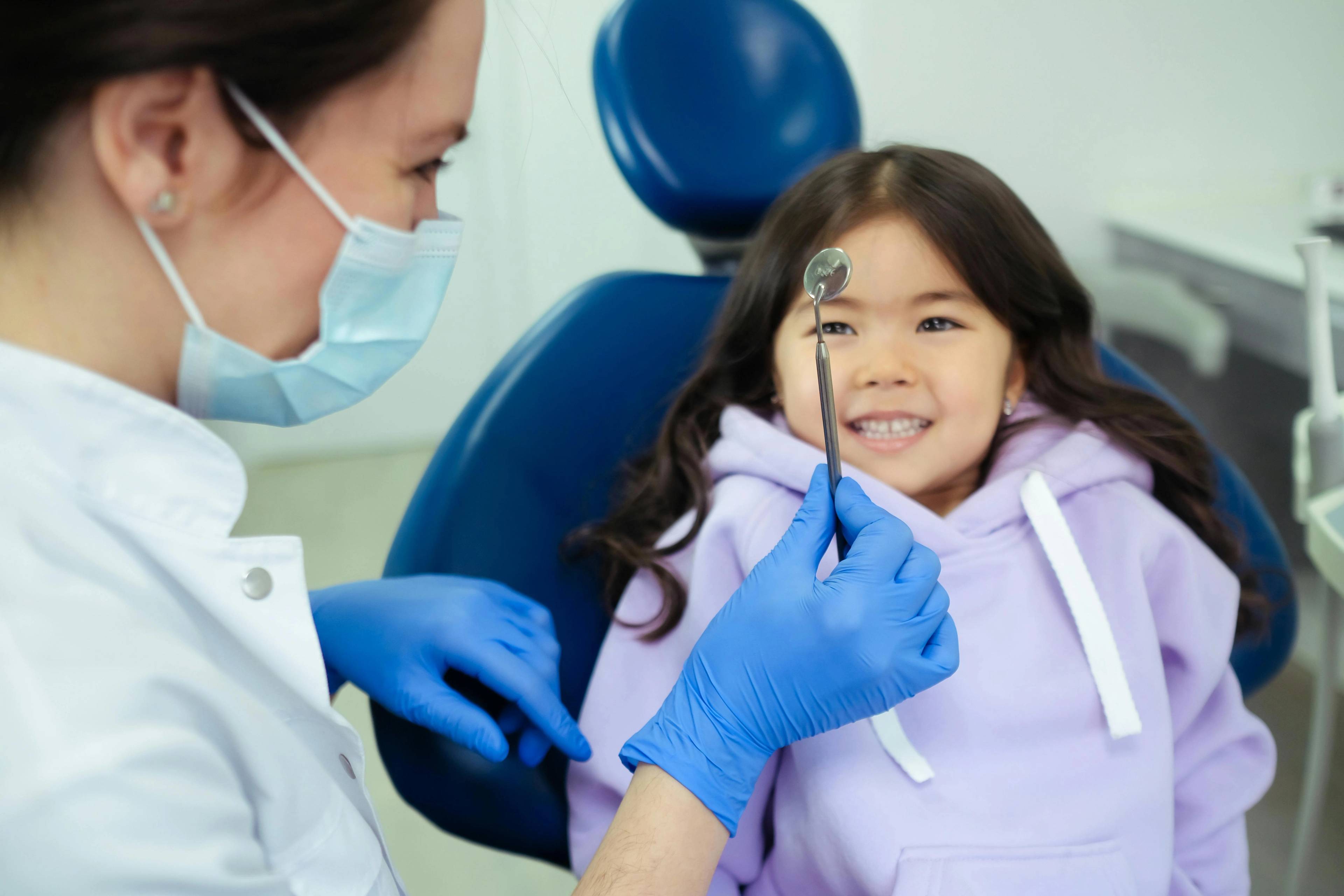 Child attending a paediatric dentistry appointment, interacting with a dentist holding a dental mirror in a friendly dental clinic environment.