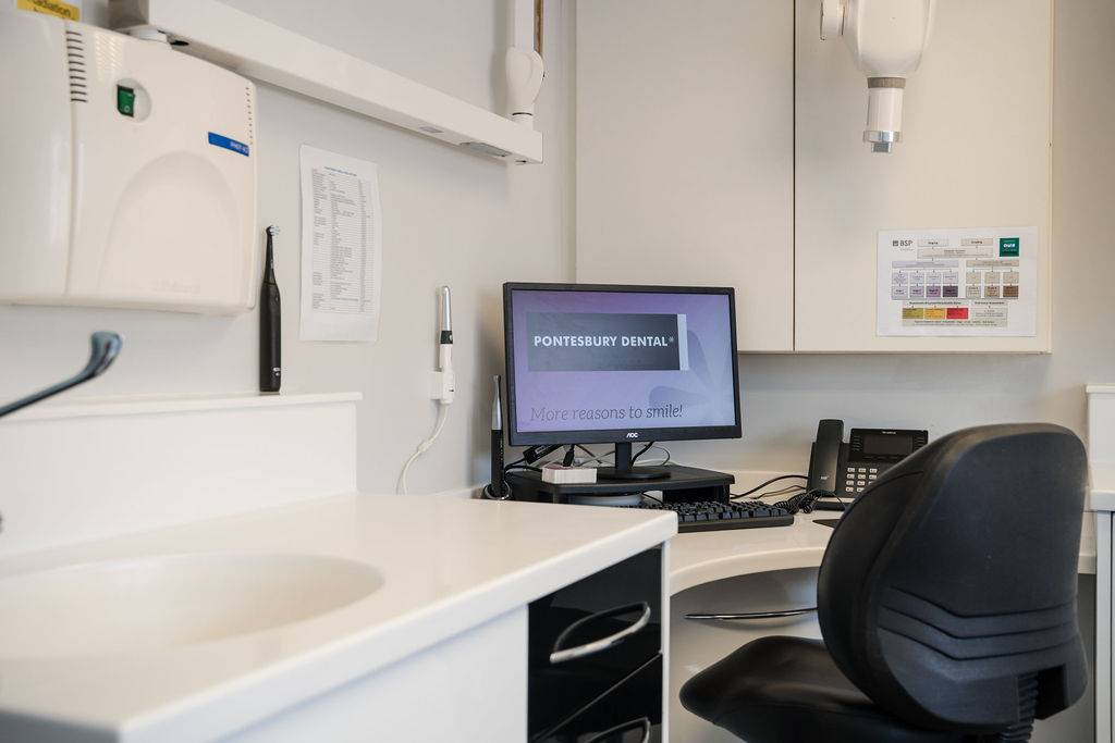 Dental treatment room with a workstation, dental equipment, and a computer monitor beside a patient examination area.