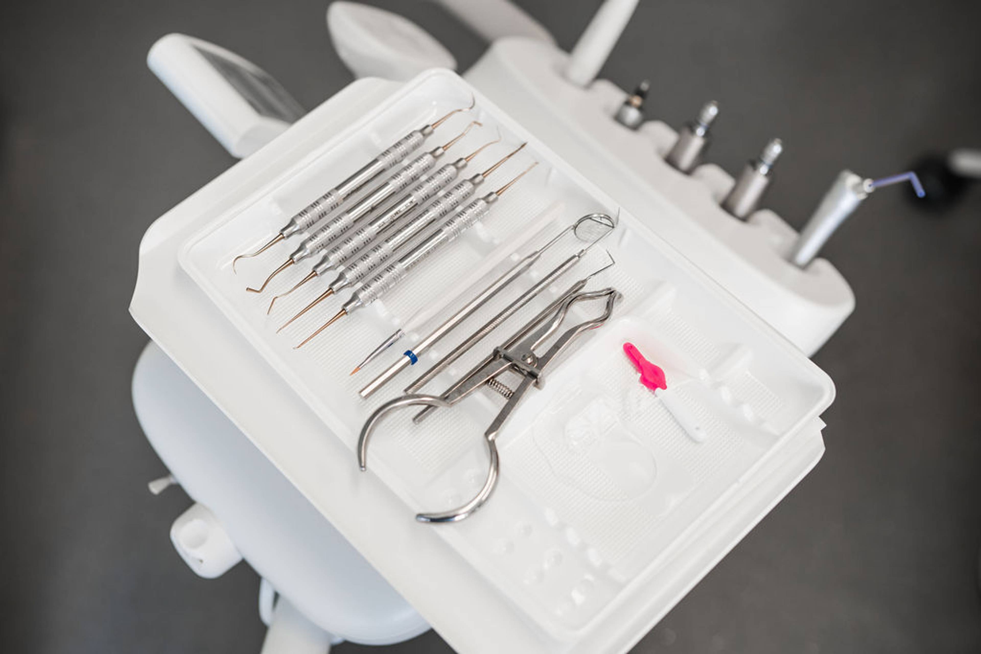 Dental instruments arranged on a tray beside a dental chair, including scalers, hand instruments, and restorative dentistry tools.