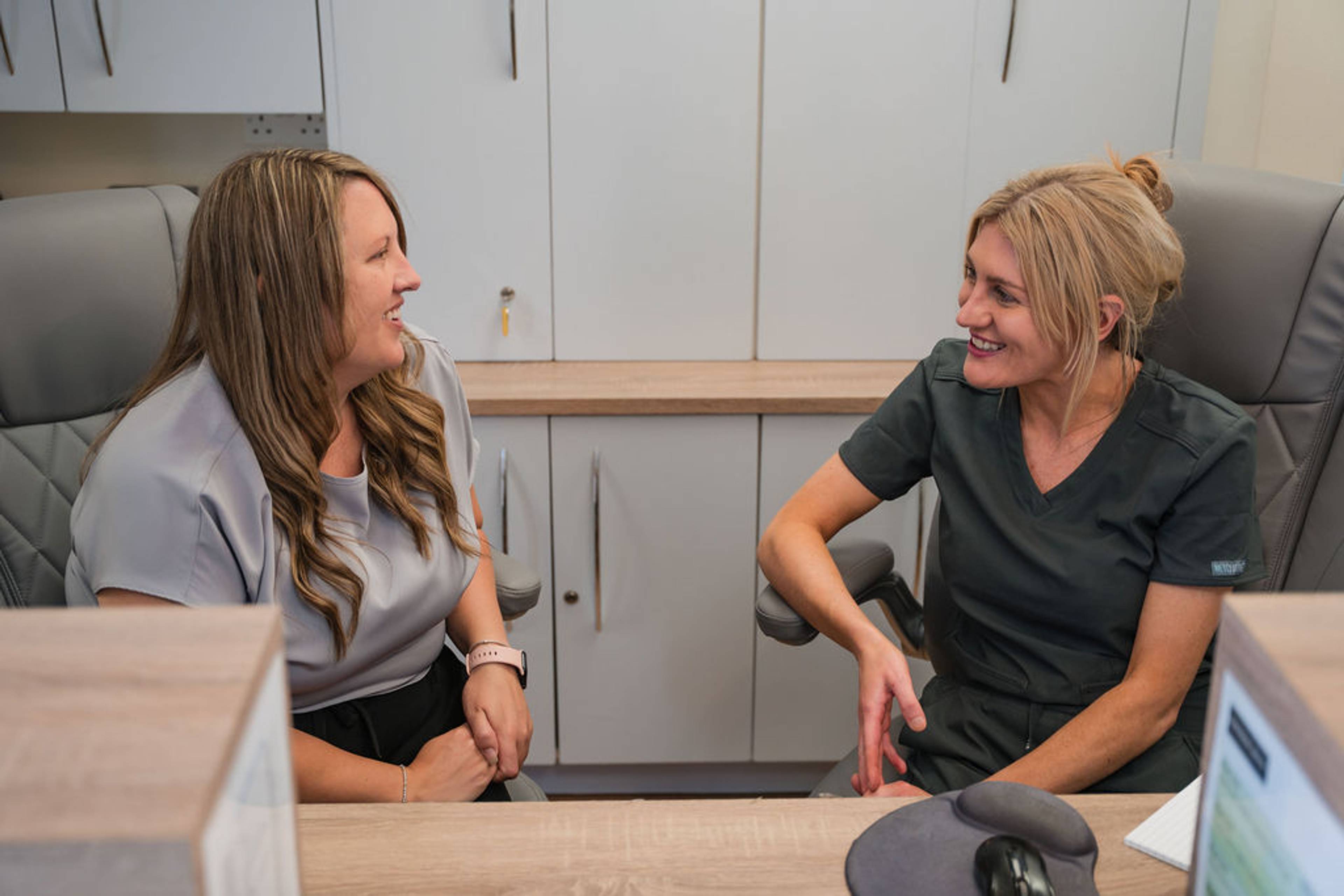 Two staff members seated at a dental practice reception desk discussing patient care and appointment coordination.
