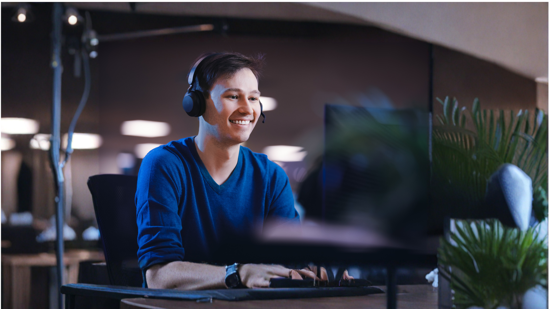 Smiling man wearing a headset with a mic, using a computer at a desk.