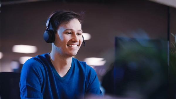 Smiling man wearing a headset with a mic, using a computer at a desk.