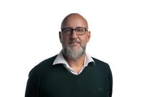 A bald man with a gray beard and glasses, wearing a sweater over a white shirt, looks at the camera.