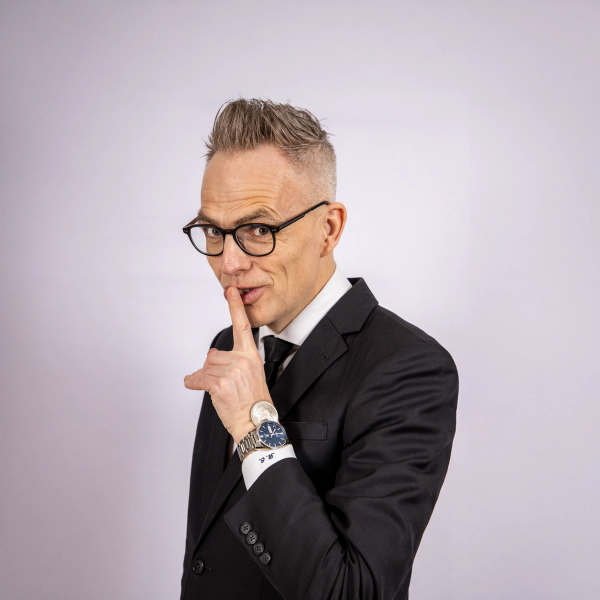 A man in a suit and glasses makes a "shhh" gesture with his finger to his lips.