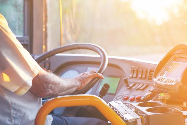 A person driving a bus, hands on the steering wheel, with sunlight streaming through the windshield.
