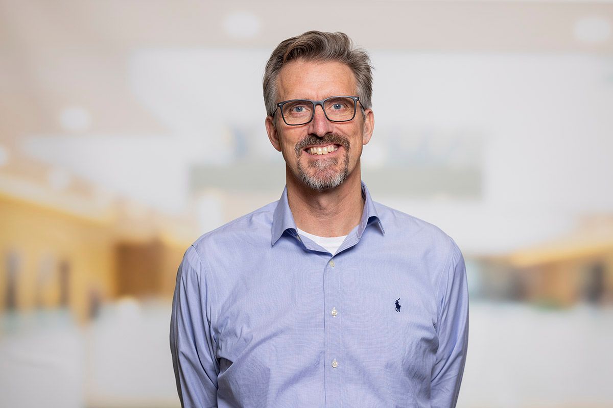 A smiling man with grey hair, a goatee, and glasses, wearing a blue collared shirt.