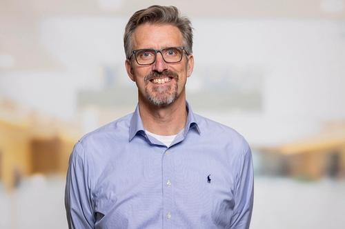 A smiling man with grey hair, a goatee, and glasses, wearing a blue collared shirt.