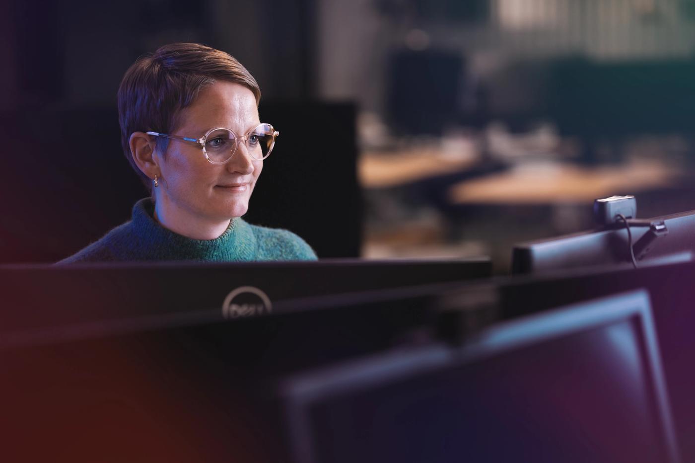 A smiling woman with short hair and glasses sits at a desk with multiple computer monitors.