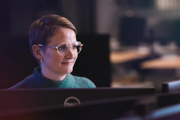 A woman with short hair and glasses smiles while looking at a computer monitor in an office.