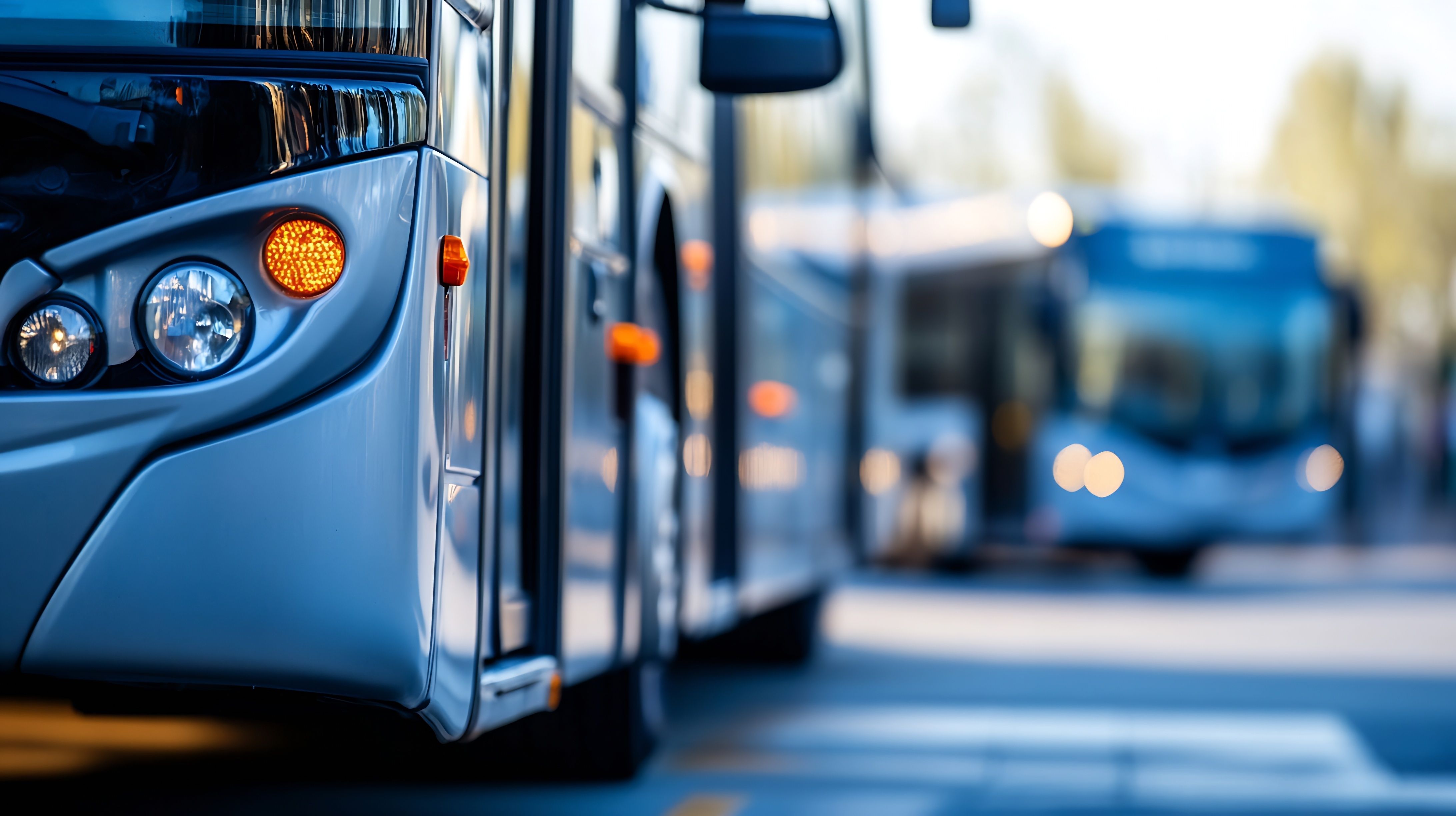 A silver bus with an orange turn signal illuminated, another bus blurred in the background.