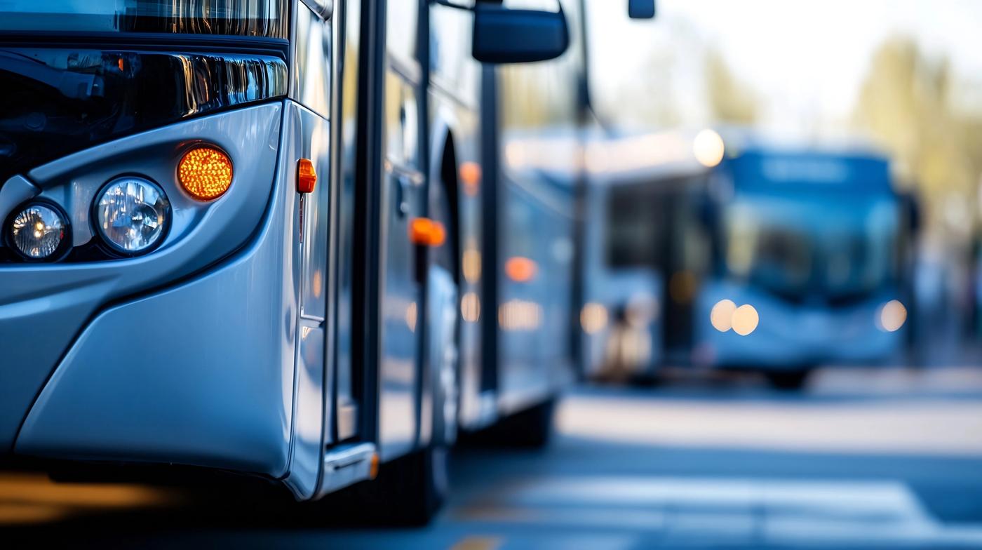 A silver bus with an orange turn signal illuminated, another bus blurred in the background.