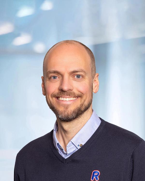 Smiling man with a beard, wearing a dark blue V-neck sweater over a light blue collared shirt.