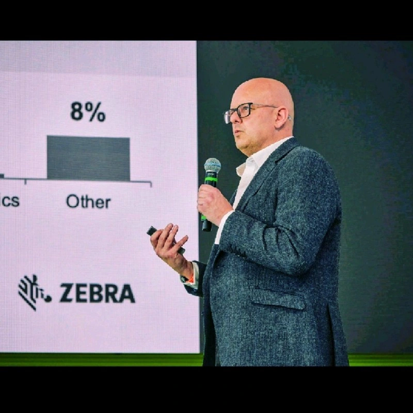 Man in glasses and a gray jacket speaking into a microphone in front of a screen displaying a bar chart with "8%" and the Zebra logo.