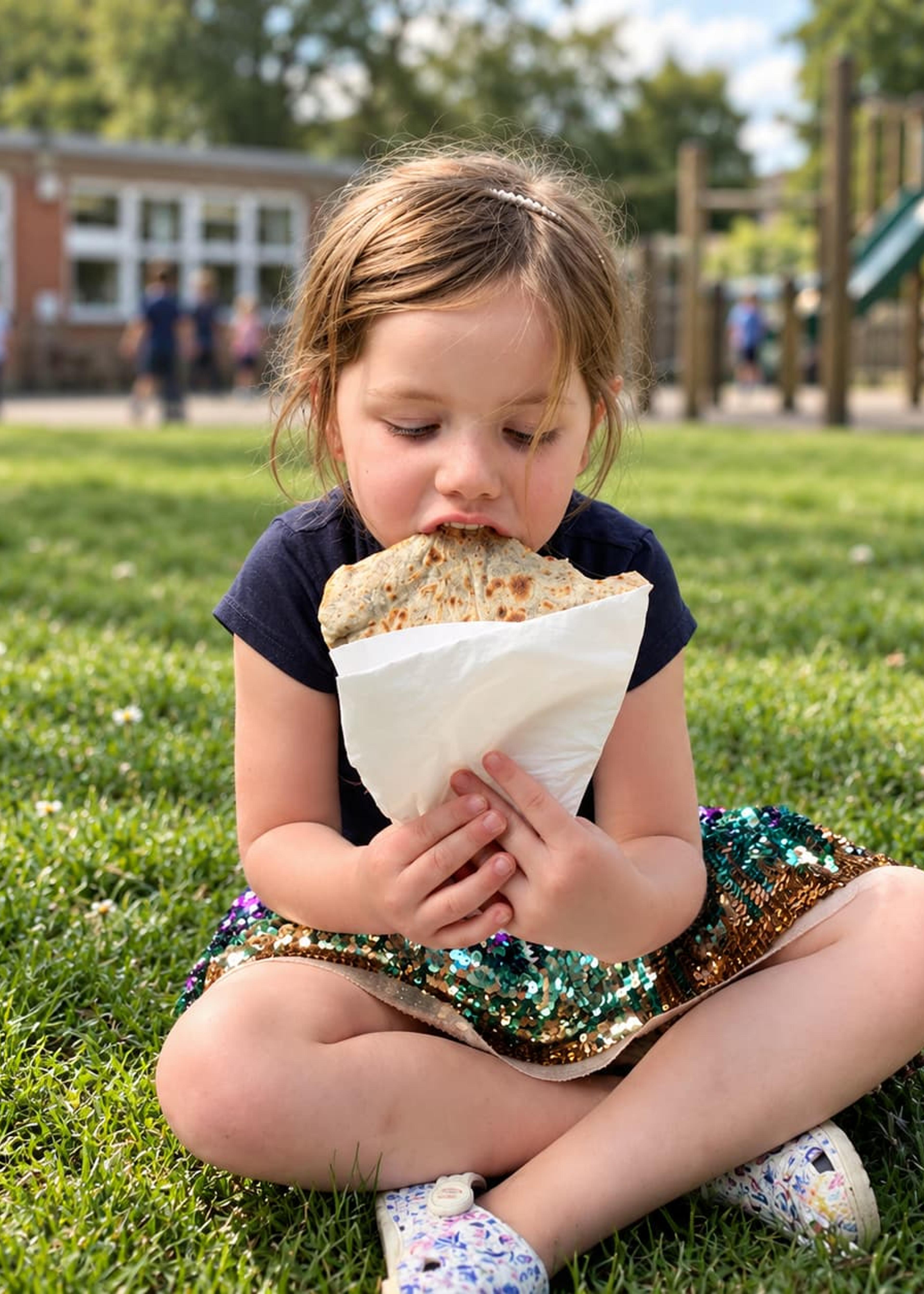 A child eating a crepe on the grass beside a playground