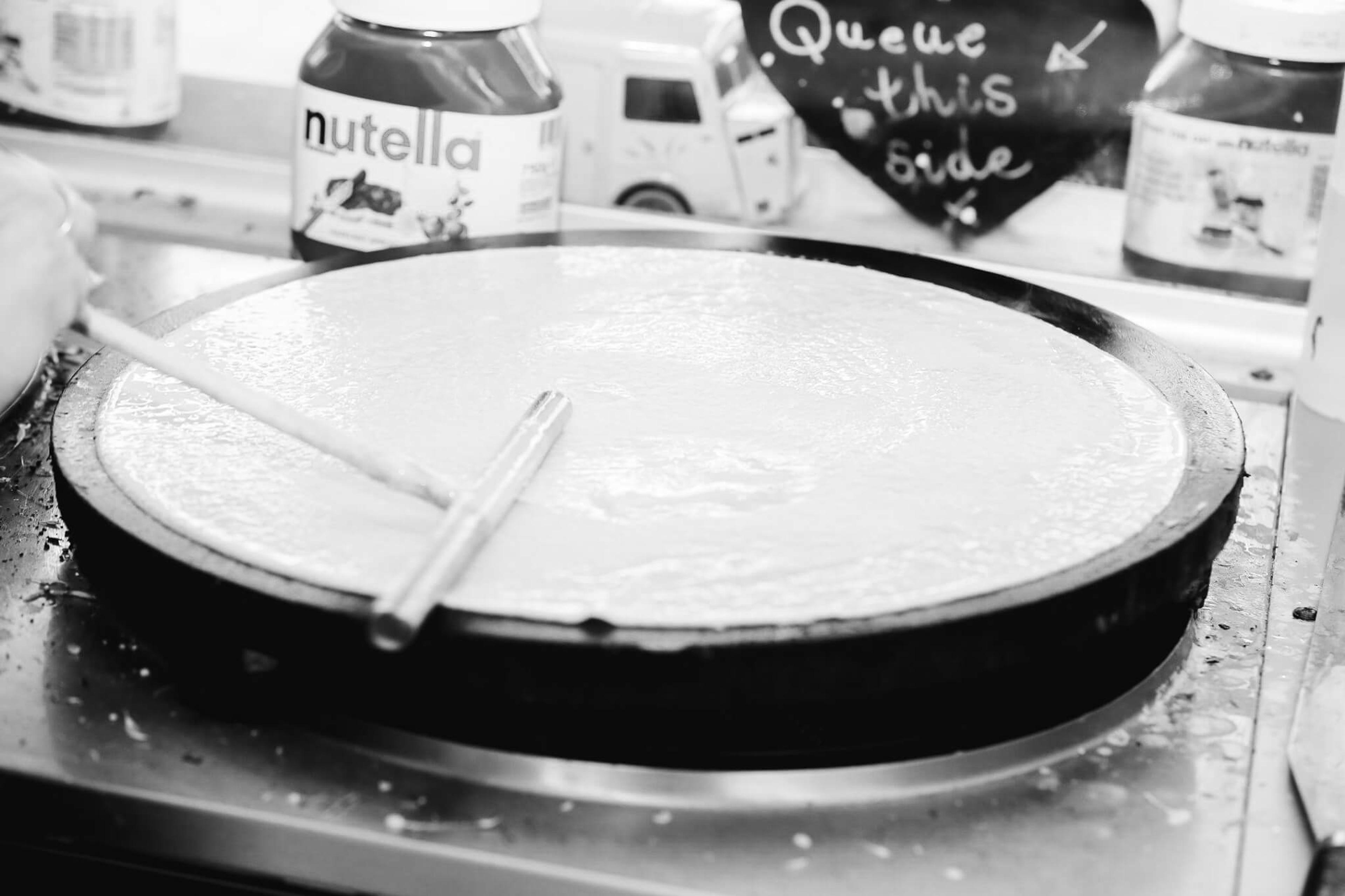 A crepe cooking on the griddle beside a jar of Nutella