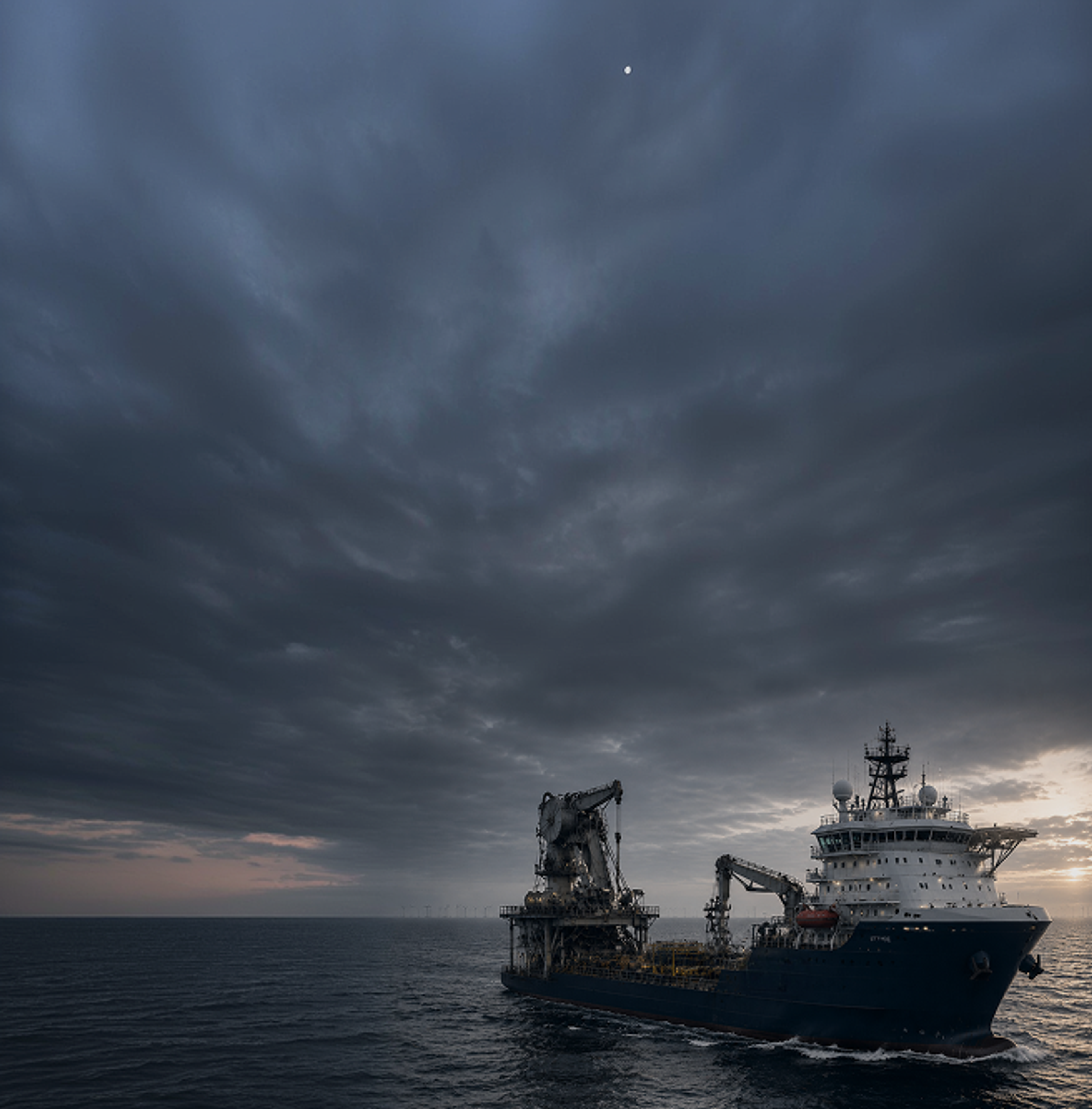 An offshore support and cable-laying vessel at sea at twilight, with wind turbines on the horizon