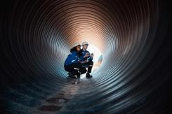 Man and a woman inside a large circular metal pipe inspecting the integrity of it